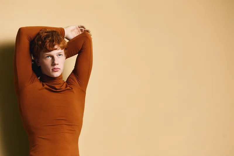 Young man with red hair wearing brown turtleneck stretching arms above head standing against beige background with soft shadows and thoughtful expression.