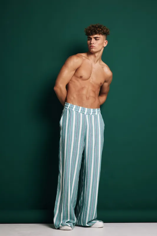 Young man standing shirtless against green background wearing striped loose pants and white shoes with curly hair and arms behind back with serious look.