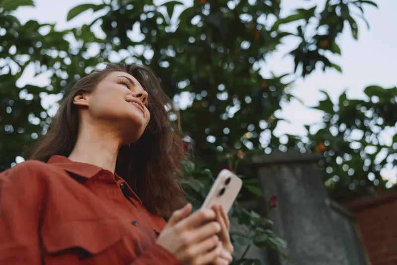 Relaxed woman outdoors holds a smartphone and looks upward, wearing a red shirt, surrounded by greenery. A lifestyle moment that conveys connection, curiosity, and everyday technology use in bright