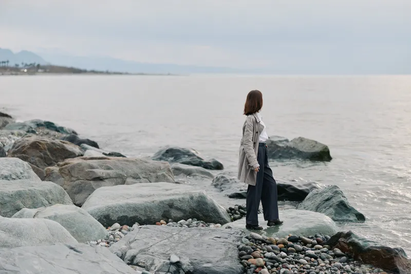 Woman in casual clothes stands on rocky shore looking at calm sea under cloudy sky. Concept of solitude, nature, and peaceful outdoor moment by the water.