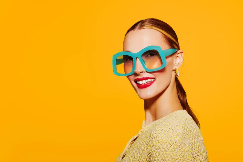 Smiling woman with large turquoise glasses and red lipstick poses against bright orange background. Fashion portrait with vibrant colors and confident expression in studio lighting.