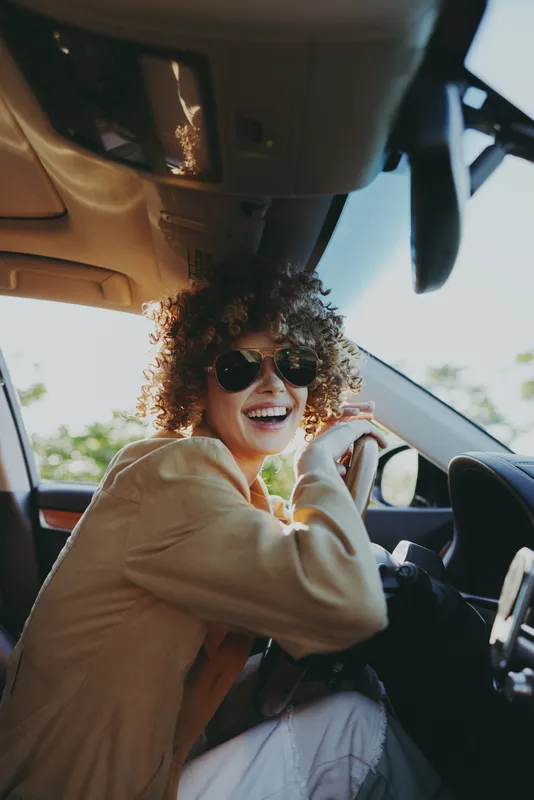 Smiling woman with curly hair driving a car, wearing sunglasses, casual jacket, warm retro film color enhances a lifestyle moment, candid and vibrant, focus on joy and motion inside the vehicle