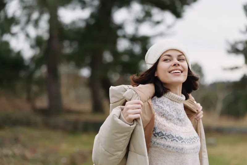 Woman outdoors wearing a cozy coat and knitted sweater, smiling in a autumn park setting, conveying comfort, style, and fresh air and energy.