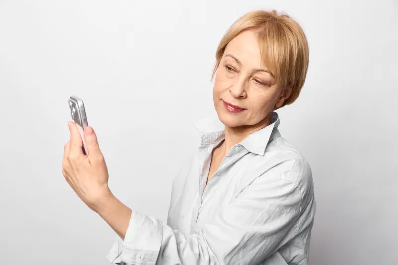 Senior woman portrait, smartphone in hand, smiling gently, indoors, white background, close up communication gesture, technology and message, calm mood, copy space