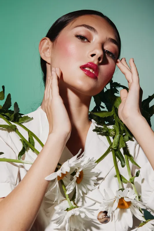 Female fashion model with glowing skin and bold red lips posing with white flowers against a green background. Creative beauty shot emphasizing makeup and floral style.