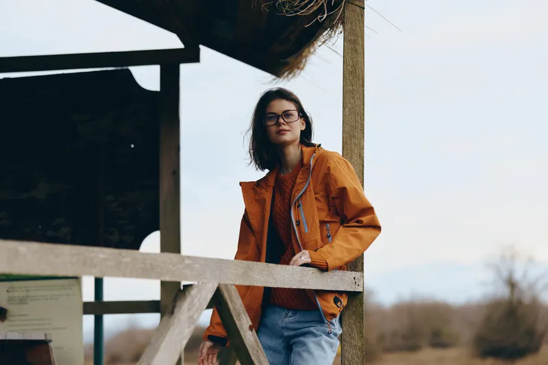 Woman lifestyle outdoors, young woman in orange jacket and glasses, standing by wooden fence, relaxed pose, looking at camera, soft wind, daytime sky, countryside field background