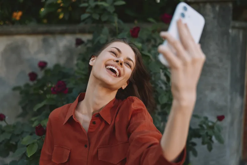 Woman lifestyle portrait taking a joyful selfie in a garden, smiling broadly, wearing a casual red shirt, natural light, candid outdoor moment among lush greenery and blooms nearby