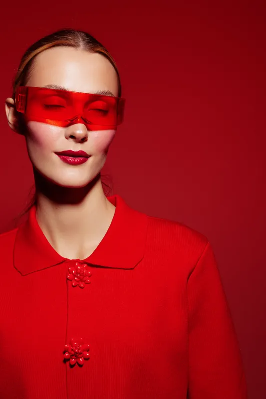 Young woman in red sweater and red translucent glasses poses with closed eyes on red background. Concept of fashion, style, monochrome, and modern portrait photography.