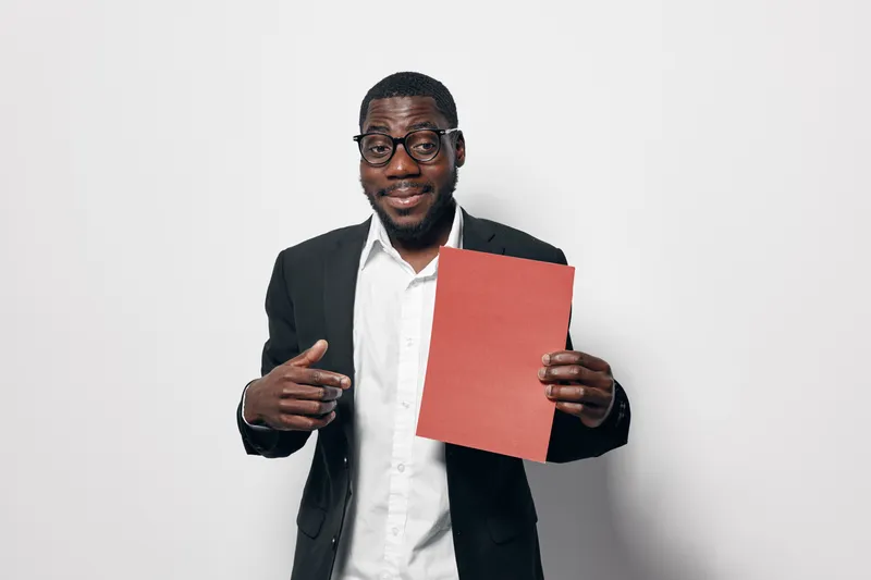 African American man with dark skin wearing glasses and black suit holding red paper and pointing at it with smile on white background.