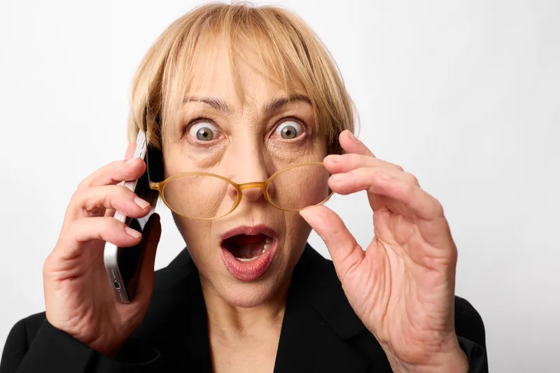 Surprised woman in studio closeup, holding eyeglasses near eyes while speaking on phone, hands up with open mouth expression, neutral background
