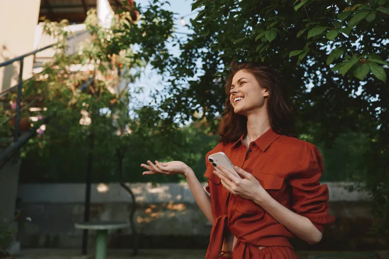 Young woman in a red dress smiles and gestures with her hand while holding a smartphone outdoors in a leafy park, capturing a candid moment of connection and everyday confidence