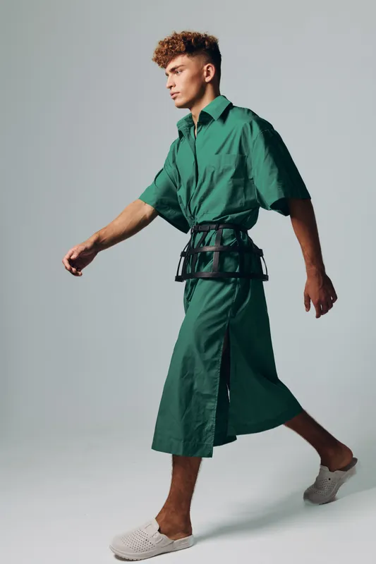 Young man walking in green oversized shirt and matching wide pants with black belt bag and white slip-on shoes on gray background in studio. Fashion, style and casual outfit concept.