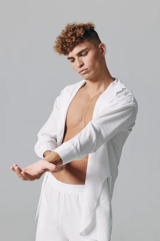 Young man with curly hair wearing white open shirt and pants adjusting sleeve in studio. Casual fashion, male model posing with bare chest and neutral background for lifestyle concept.