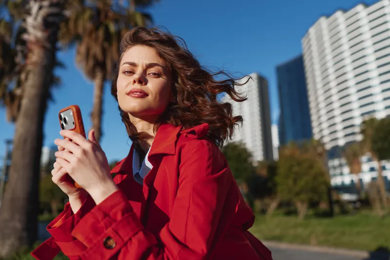 Young woman in red coat holding smartphone outdoors, confident expression, sunny day, urban park setting, modern buildings in background