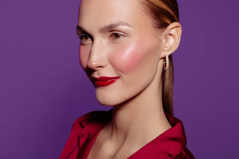 Close-up portrait of a confident woman with red lipstick and glossy skin wearing a red blouse against a purple background. Concept of beauty, makeup, fashion, and elegance.