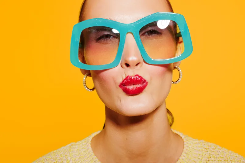 Young woman with large blue sunglasses and red lipstick makes a kiss face on bright yellow background. Fashion portrait with colorful accessories and playful expression concept.