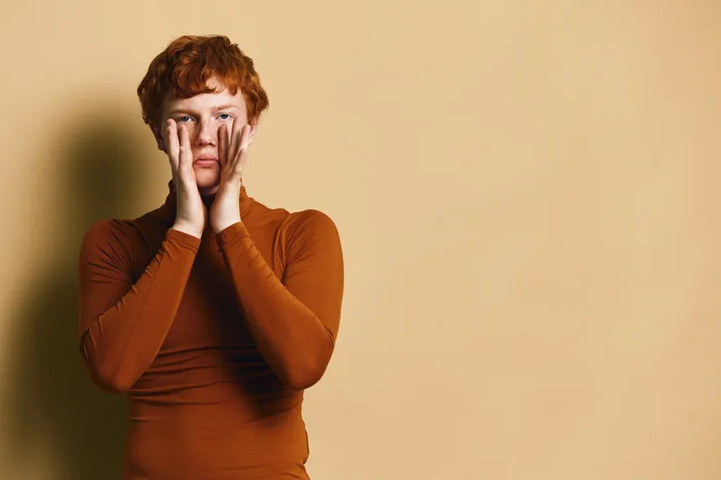 Young man with red hair touches face with hands, wearing orange shirt, standing against beige background looking thoughtful and serious in studio portrait.