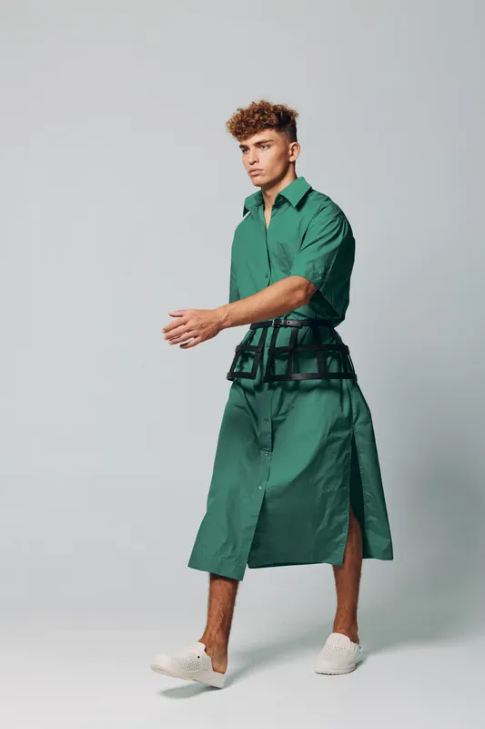 Young man with curly hair walking isolated on grey studio background wearing modern green designer outfit shorts and shirt combination creative fashion with futuristic accessories