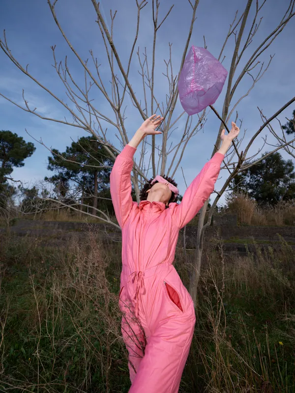 Woman in a pink jumpsuit releases a lantern outdoors among bare trees, celebrating a joyful moment with open sky and natural field surroundings.