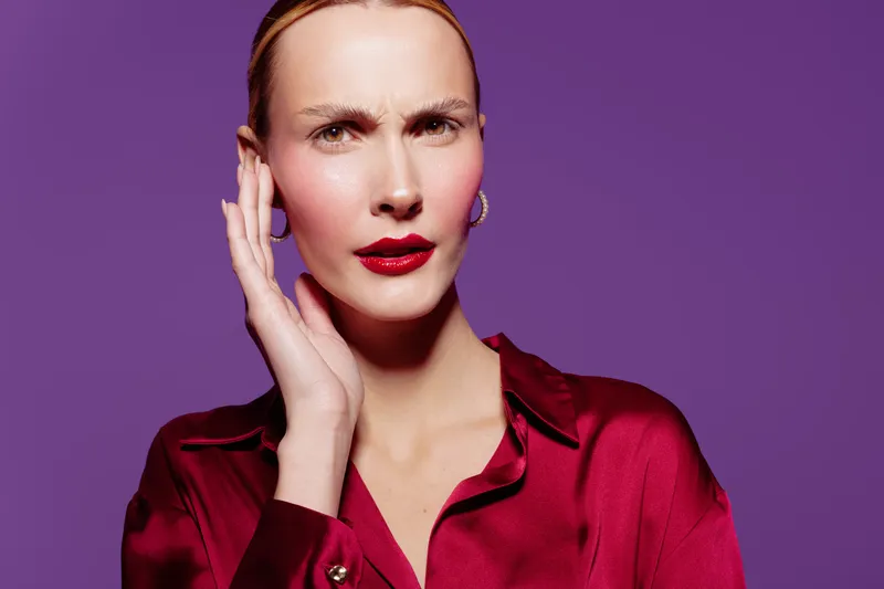 Confused woman in red satin blouse touches face with hand, looking puzzled on purple background. Concept of doubt, uncertainty, and questioning emotions in studio portrait.