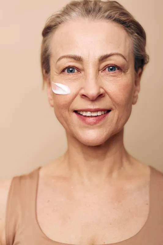 Smiling mature woman with blue eyes applies white cream on cheek in soft studio light. Concept for skincare, aging skin care, natural beauty, wellness, and facial moisturizing routine.