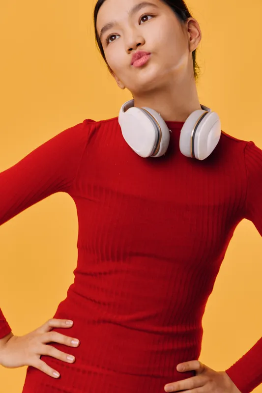 Young woman in red dress with white headphones around neck posing against yellow background, confident expression and hands on hips in studio portrait. Fashion, lifestyle and music concept.