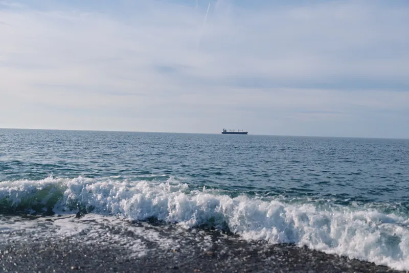 Sea scene with gentle waves breaking on a pebbled shore and a distant ship on the horizon under a clear sky, capturing calm coastal mood and open ocean.
