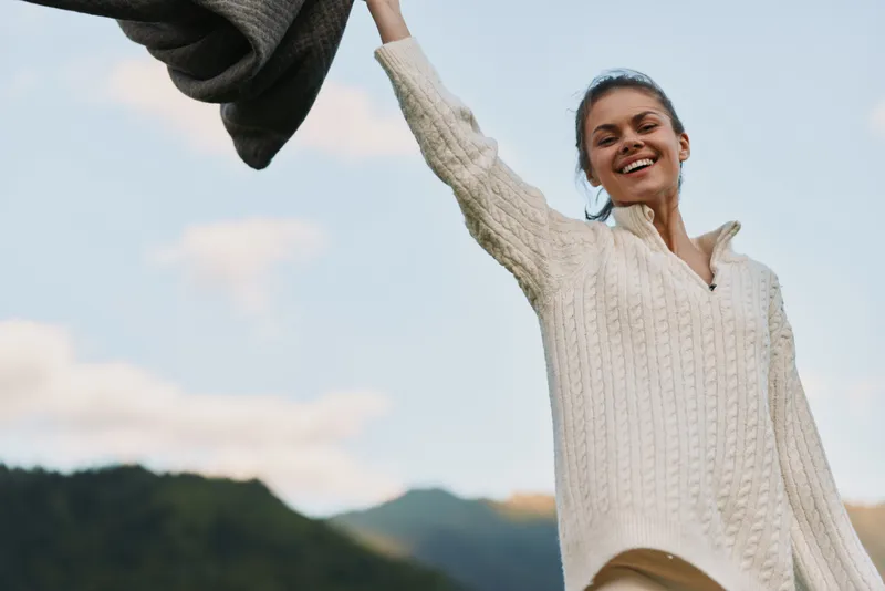 Joyful young woman in a cream knit sweater raises her arm outdoors, smiling against a blue sky and distant hills.