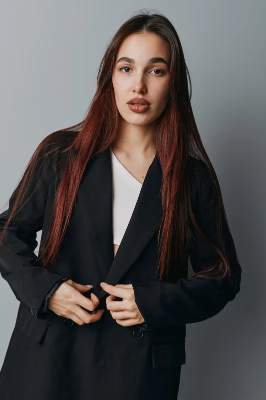 Young woman with long hair wearing black blazer and white top posing against gray background in studio. Portrait of confident female model with natural makeup and serious expression.