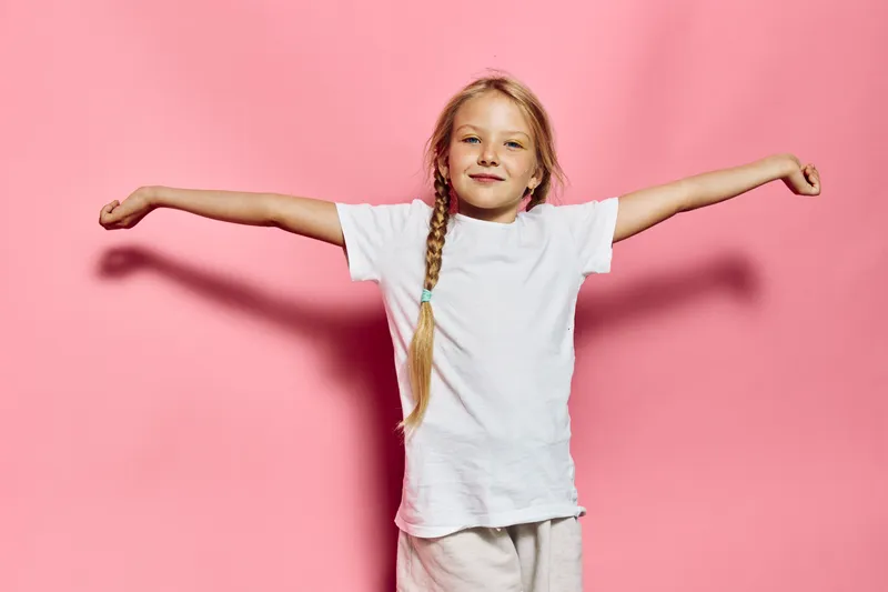 Happy young girl with long braided hair stands with arms outstretched against a pink background. She wears a white t-shirt and smiles gently.