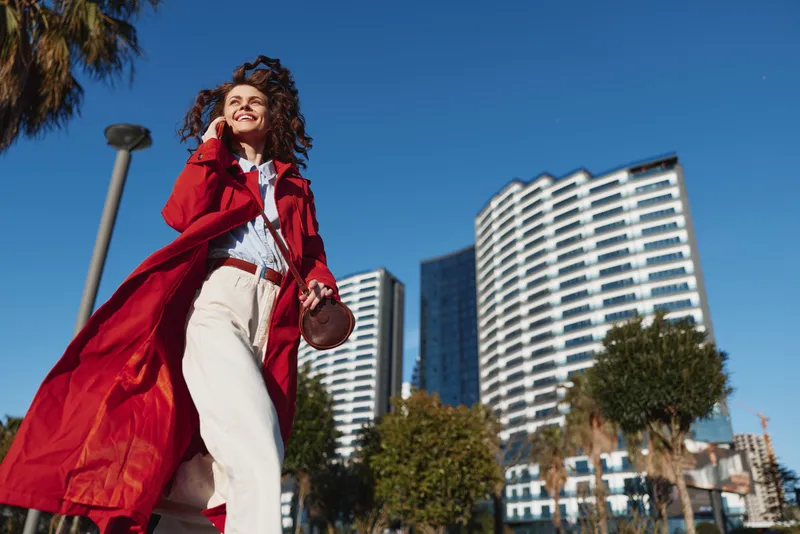 Smiling woman in a red coat walking outdoors among modern buildings. Urban street fashion vibe with bright daylight and a confident, carefree mood and glow.