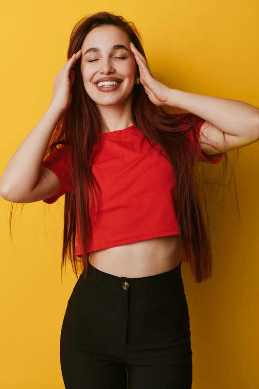 Young woman with long hair smiling with closed eyes and hands near face wearing red crop top and black pants standing against yellow background in studio.
