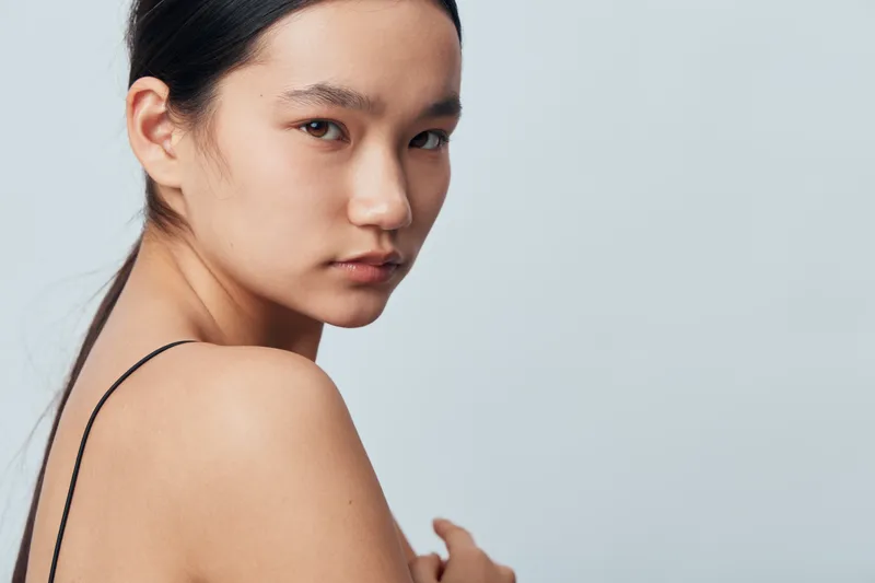 Portrait of young woman with clear skin and natural makeup looking over shoulder with serious expression. Minimalist style with bare shoulder and simple background in studio lighting.
