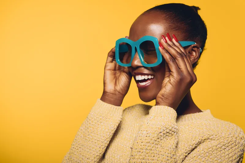 Smiling dark skinned African woman with natural hair wearing oversized blue sunglasses and beige sweater on yellow background. Concept of joy, fashion, and vibrant lifestyle.