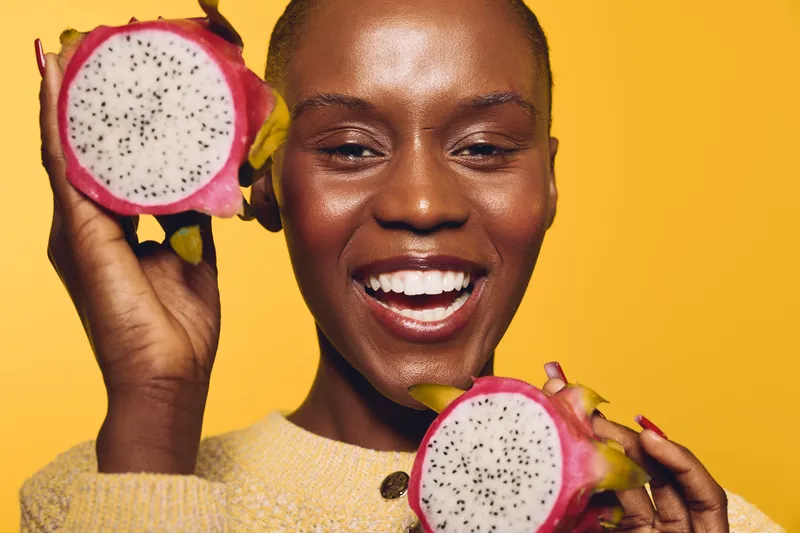 Happy African woman with dark skin holding two halves of dragon fruit near her face, smiling on yellow background. Concept of healthy lifestyle, tropical fruit, and natural beauty.