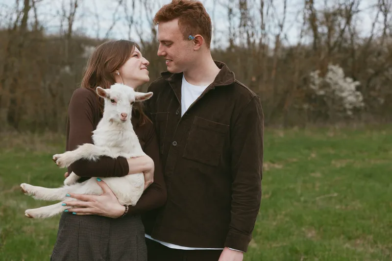 A couple outdoors in a grassy field cradle a small goat kid, smiling as they look at each other. A warm lifestyle moment showcasing affection, rural life, and gentle bonding with animals in a bright