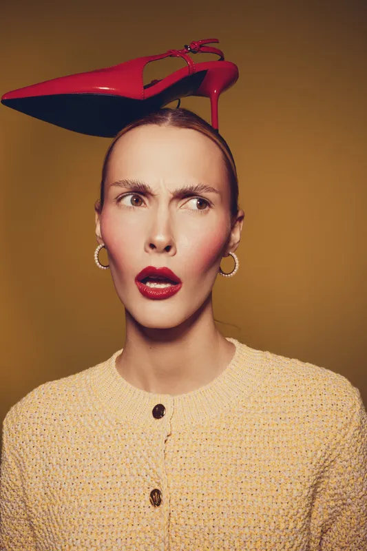 Confused woman with red lipstick and hoop earrings balances red high heel shoe on her head. Yellow background and beige textured sweater create a stylish and quirky fashion portrait.