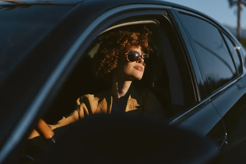 Smiling woman with curly hair sits in a car, wearing sunglasses, bathed in warm golden hour light. Retro film color enhances a confident, stylish mood of urban travel and self expression.
