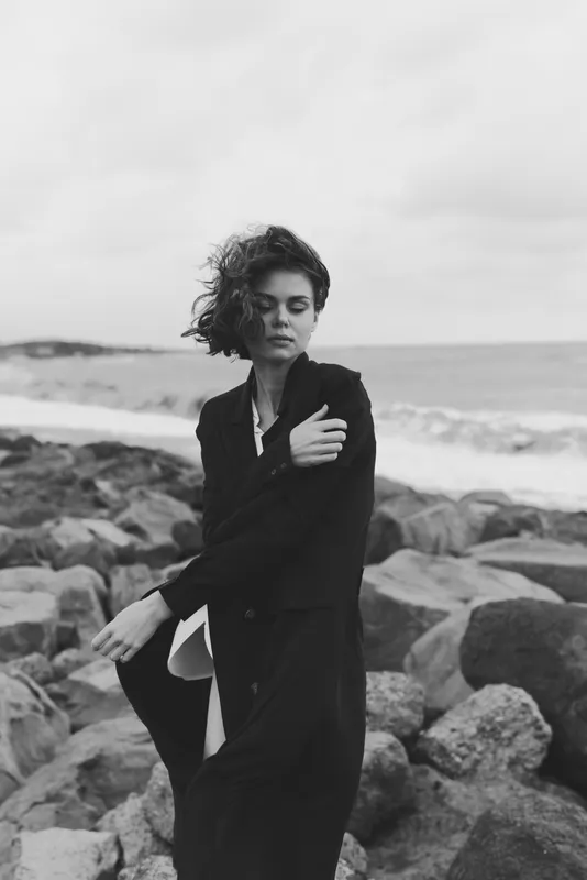 Coast, windy fashion, black and white silhouette by the seaside on rocks, windswept mood with an elegant coat, moody ocean breeze, dramatic monochrome storytelling
