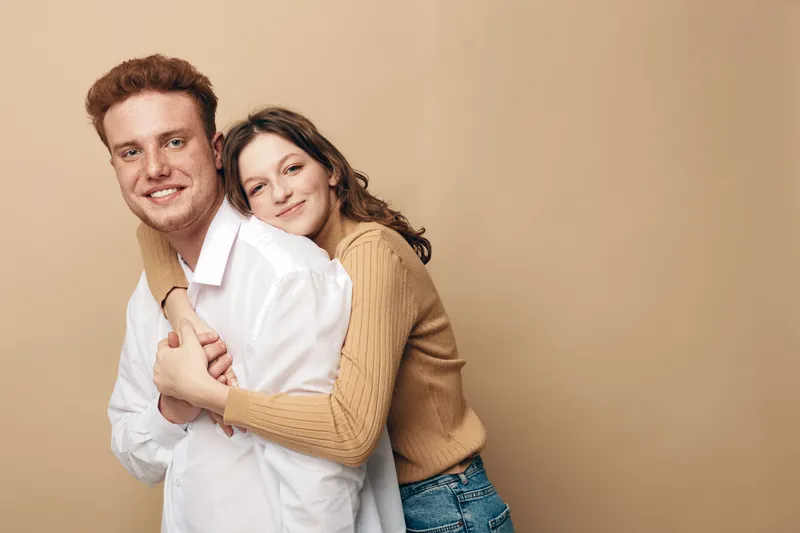Smiling young couple embraces in casual clothes against beige background. Happy man and woman show affection and warmth, concept of love, relationship, friendship, and togetherness.