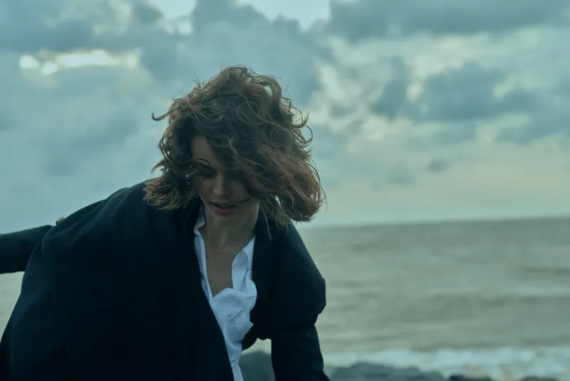 Windy coastal scene with a woman in a dark blazer moving along the shore, hair blown by gusts, focused expression and moody sky, conveying determination, resilience and professional energy by the sea.
