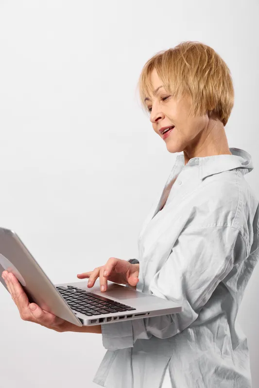 Senior woman with laptop typing in a white indoor scene, technology work and communication for remote online productivity and focus, professional career planning during study