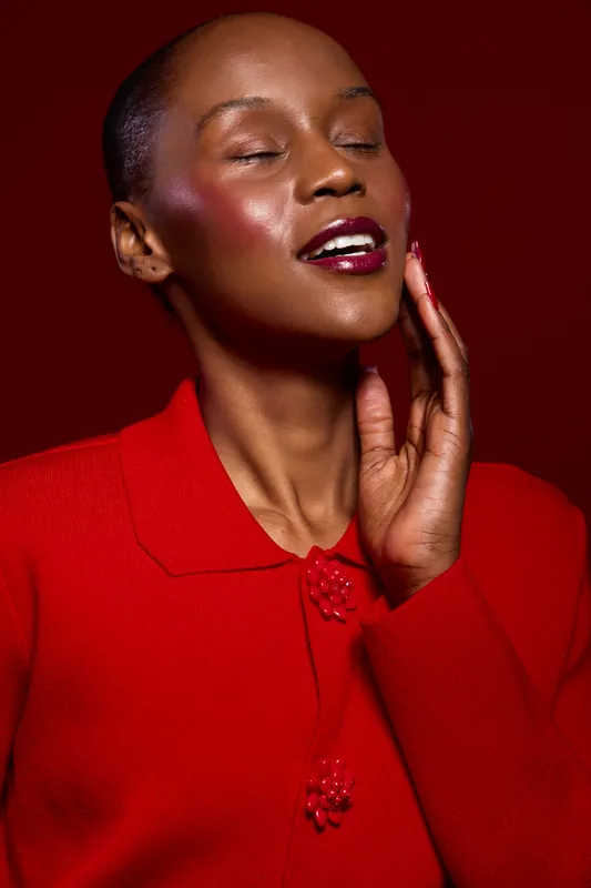 Portrait of a happy black woman with dark skin wearing a red sweater against a red background. She smiles with closed eyes and touches her face, expressing joy and confidence.