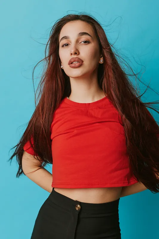 Young woman with long hair wearing red shirt and black pants posing confidently against blue background. Female model with natural makeup and casual style in studio portrait.