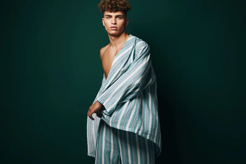 Young man with curly hair wearing striped pajamas posing against dark green background. Fashion portrait with casual style and confident expression in studio lighting.