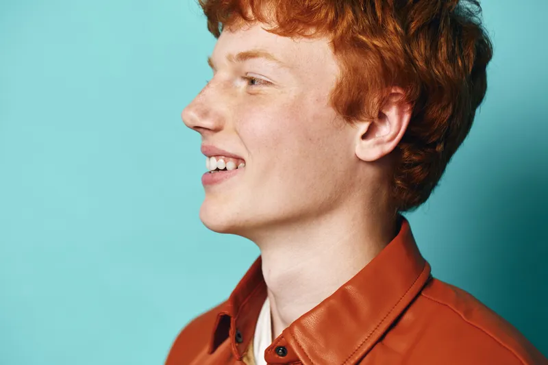 Young man with red hair wearing orange jacket smiling and looking sideways against blue background. Profile portrait of happy teenager with casual outfit and natural expression.