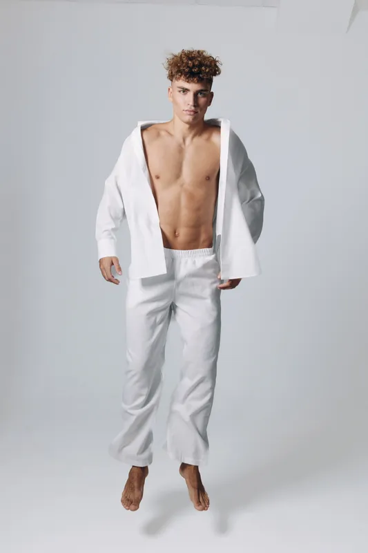 Young man with curly hair wearing white pants and open white shirt standing barefoot on gray background in studio. Male model posing with confident expression and casual style.