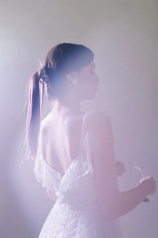 Young woman in a white lace dress with a ponytail stands in soft light, looking to the side. Concept of beauty, fashion, elegance, and delicate femininity in a serene atmosphere.