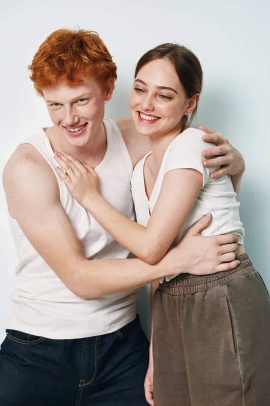 Young couple smiling and embracing in casual clothes standing close together against white background. Happy redhead man and brunette woman in relaxed pose with affection and joy.