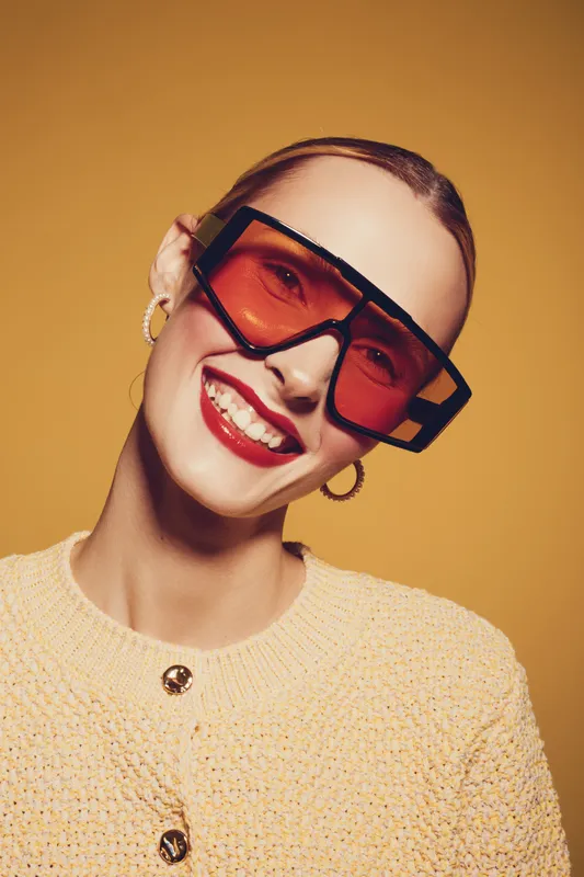 Smiling woman with red lipstick wearing large orange sunglasses and hoop earrings poses against yellow background. Fashion portrait with bright colors and joyful mood.